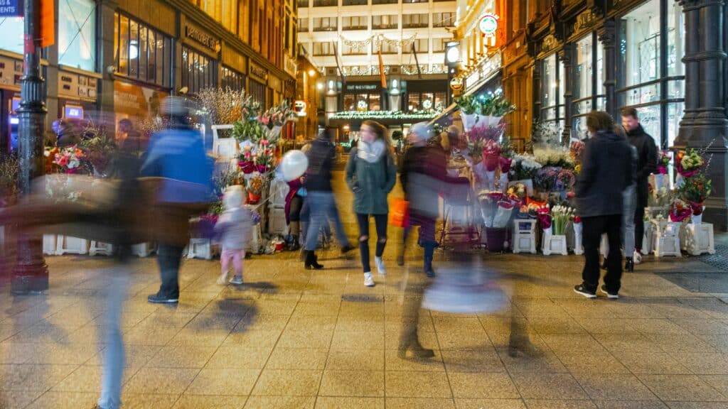 People walking through a busy, decorated retail street at night with holiday shopping activity and flower stands along the sidewalk.